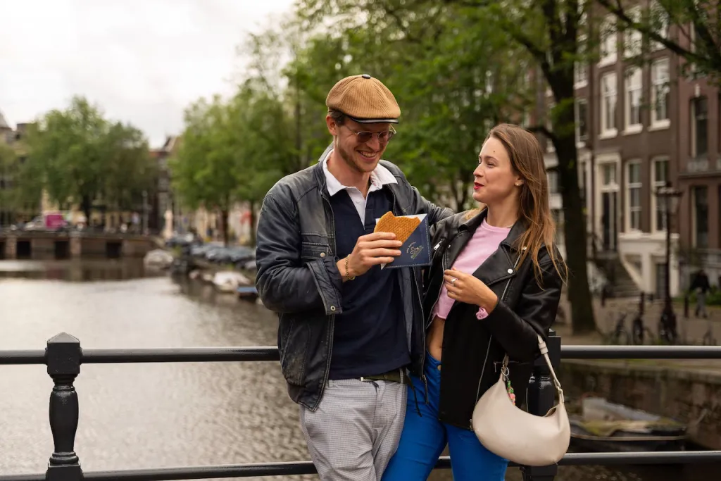 Smiling couple shares a fresh stroopwafel in a blue paper sleeve while leaning on a canal bridge railing in Amsterdam