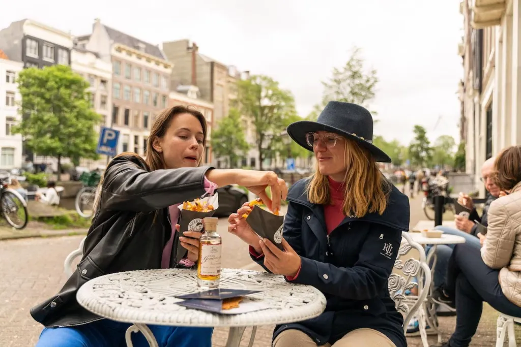 Two women at a white terrace table share cones of fries topped with red onion, with Amsterdam canal houses in the background