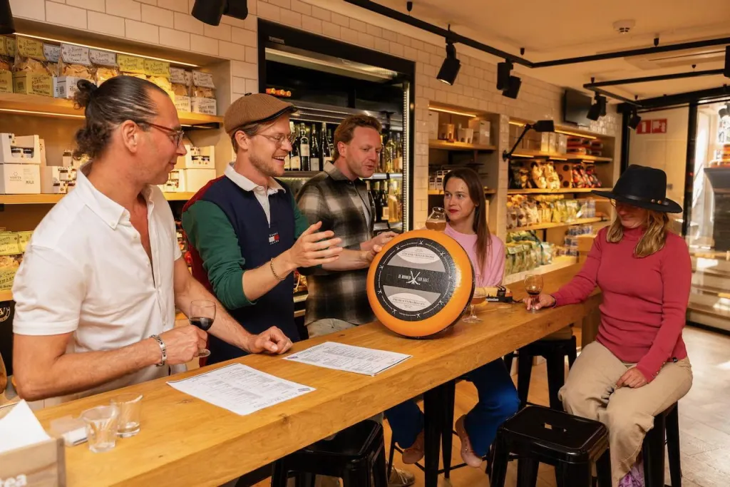 Shopkeeper presents a whole cheese wheel to four guests seated at a long wooden counter in an Amsterdam specialty food shop