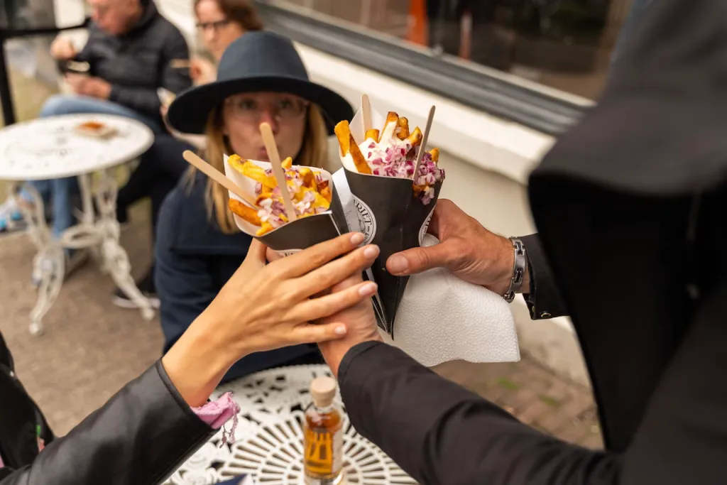 Hands clink two paper cones of fries topped with red onion together over a terrace table while a woman in a hat watches