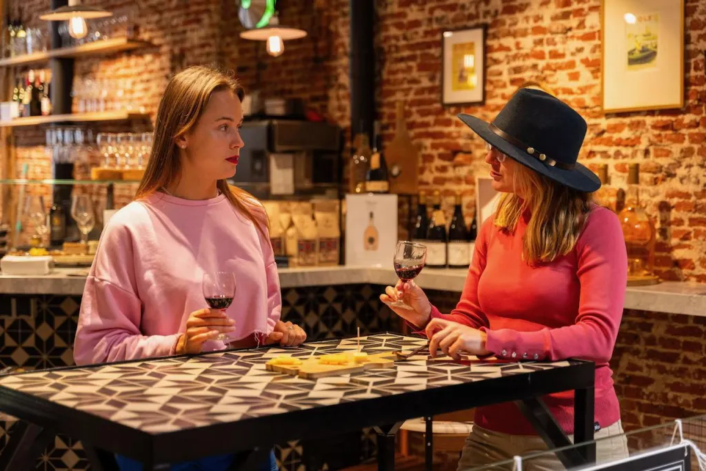 Two women talk over glasses of red wine and a small board of cheese at a tiled table in a brick-walled Amsterdam wine bar