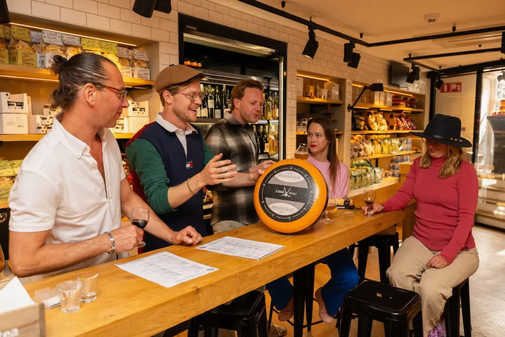 A guide holds up a whole wheel of Gouda cheese while talking to a small group seated at a wooden counter in an Amsterdam cheese shop