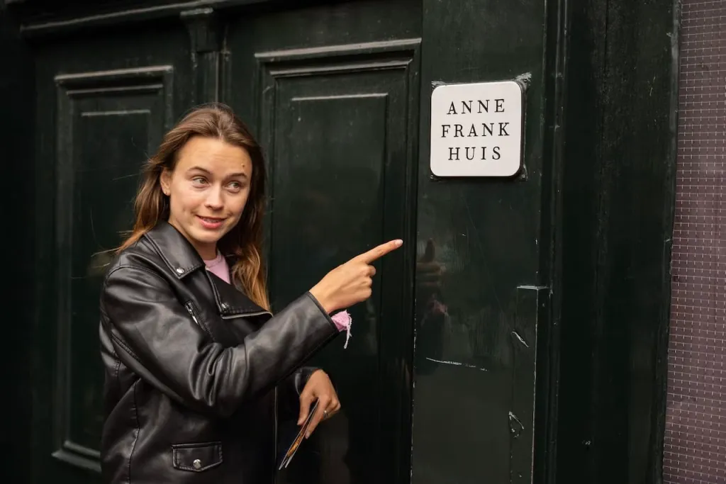 A smiling woman in a leather jacket points at the Anne Frank Huis sign on a dark green wooden door in Amsterdam
