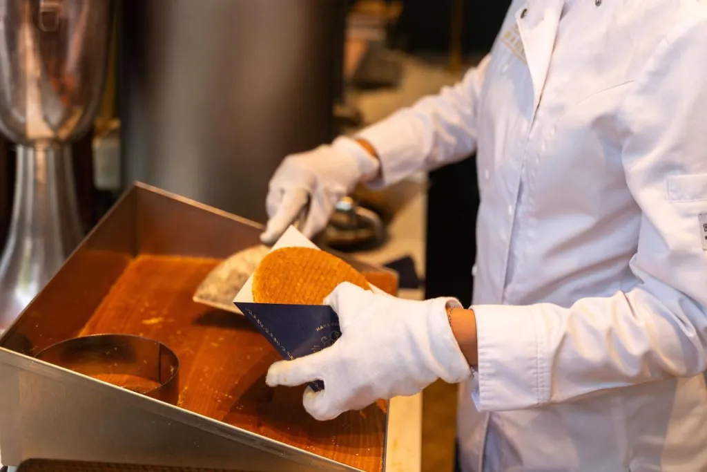A baker in a white coat and gloves slides a freshly made stroopwafel into a paper sleeve at the counter