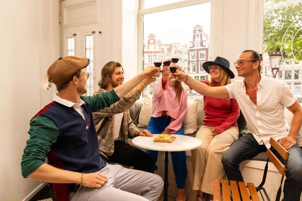 Five people raise glasses of red wine around a small marble table by a bright window with Amsterdam canal houses outside