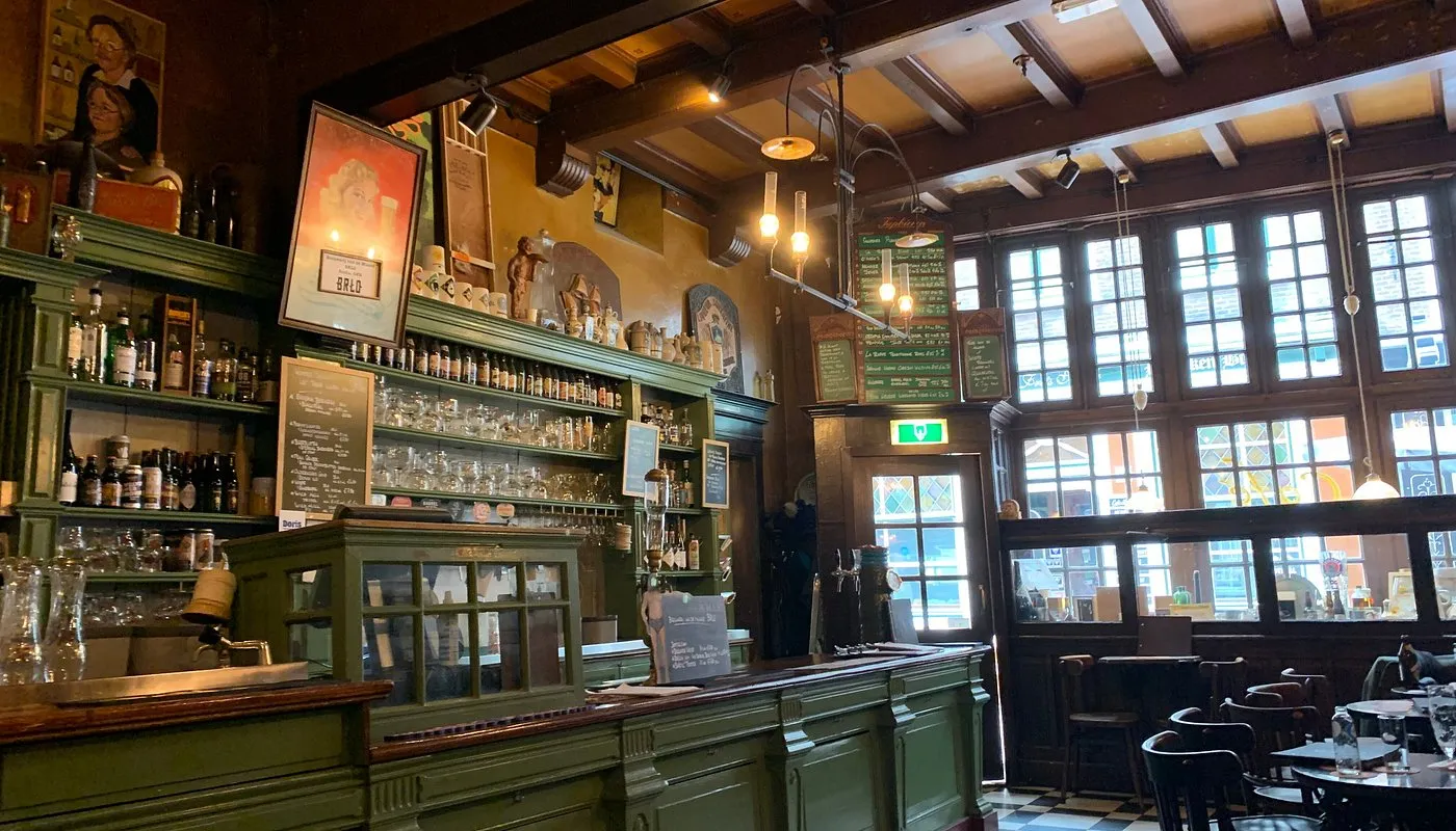 Interior of a historic Amsterdam bar with a green wooden bar, shelves of bottles and glasses, chalkboard beer menus, and a high beamed ceiling