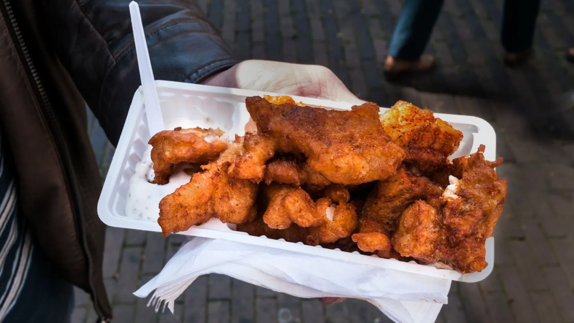 A hand holds a white tray piled with golden battered kibbeling fish bites and garlic sauce on a street