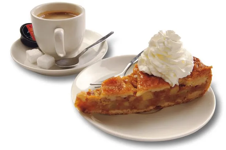 A slice of Dutch apple pie topped with whipped cream beside a cup of coffee with sugar cubes on a white background