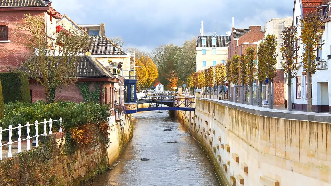 A narrow stream runs between brick houses and autumn trees in a small Dutch town, crossed by a blue footbridge
