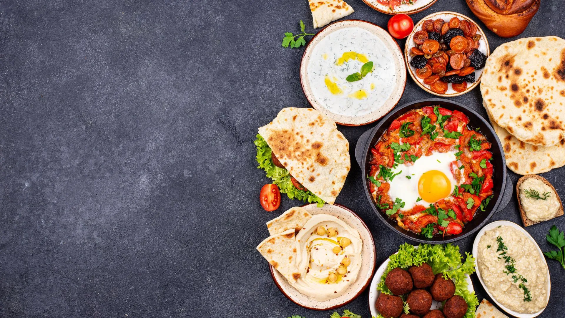 Overhead view of Middle Eastern dishes including shakshuka, hummus, falafel, and flatbreads arranged on a dark surface