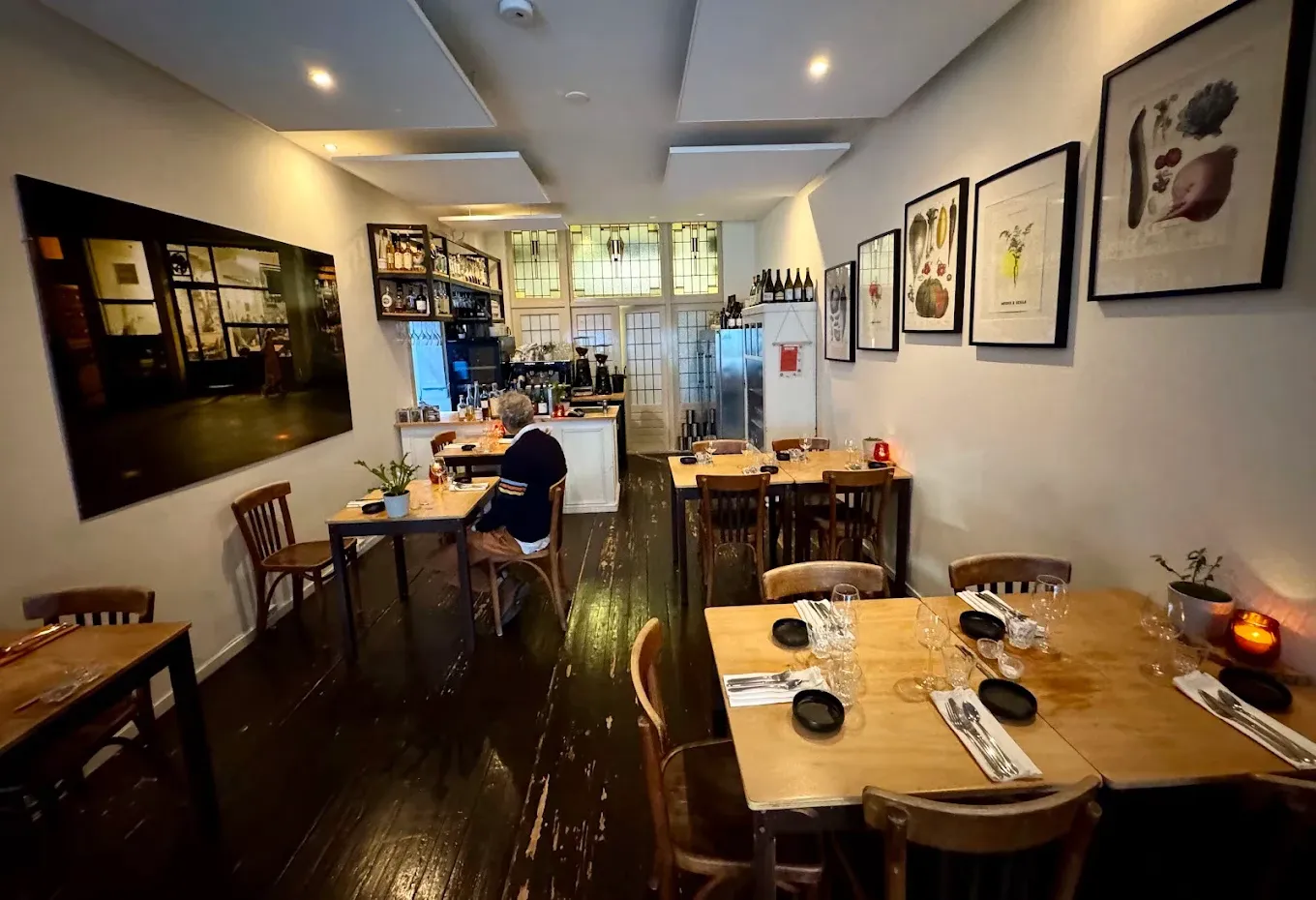 A small restaurant interior with wooden tables set with cutlery and wine glasses, botanical prints on white walls, and a dark wooden floor