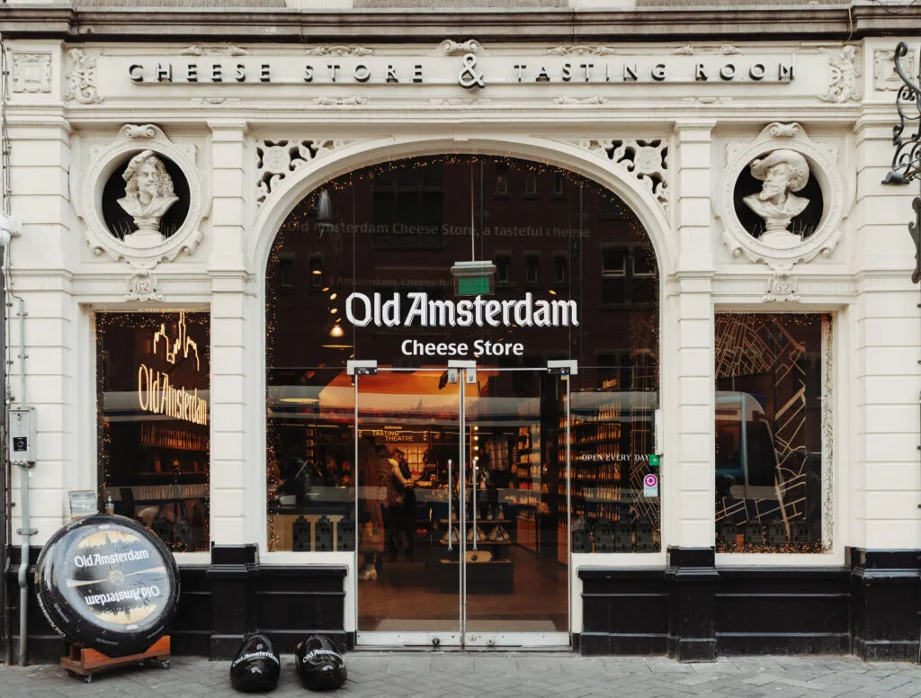 Ornate white facade of the Old Amsterdam Cheese Store and Tasting Room with a black cheese wheel barrel and wooden clogs by the entrance