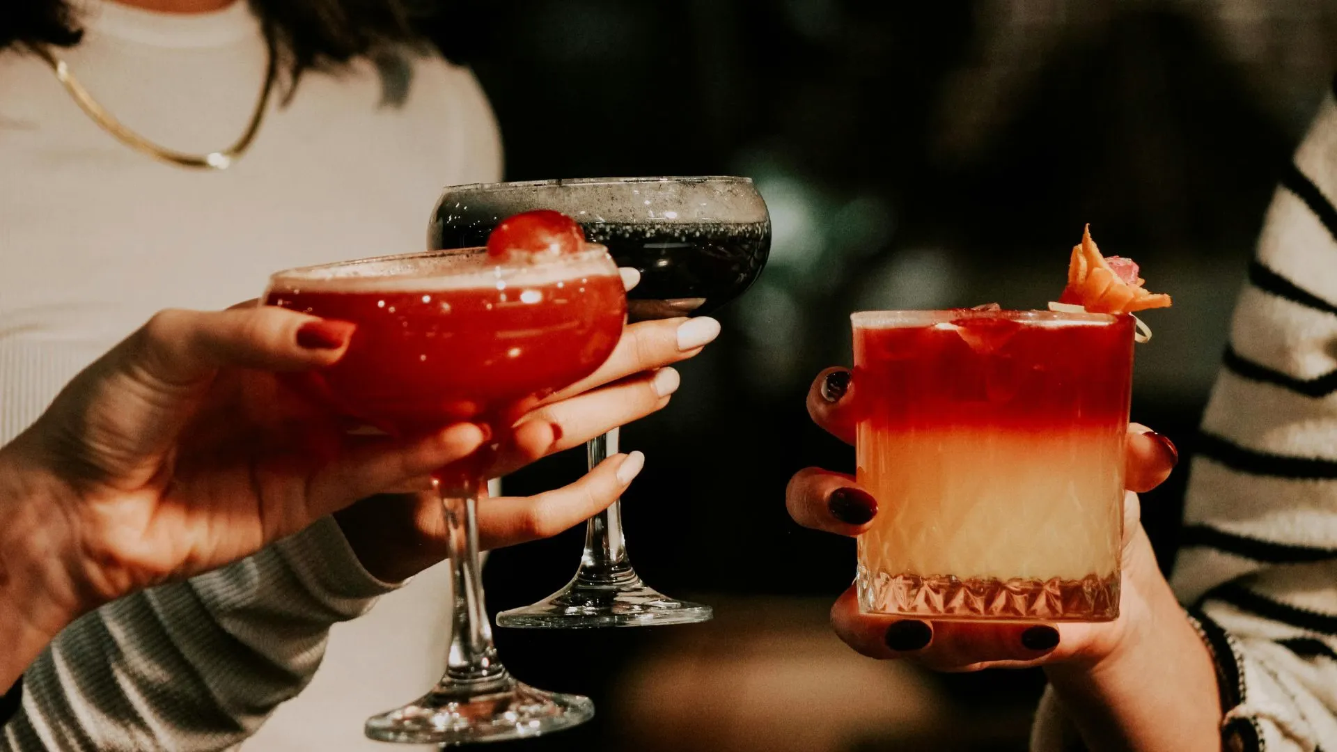 Three hands clinking colorful cocktails, a red coupe, a dark coupe and a layered red and yellow drink, in a dimly lit bar