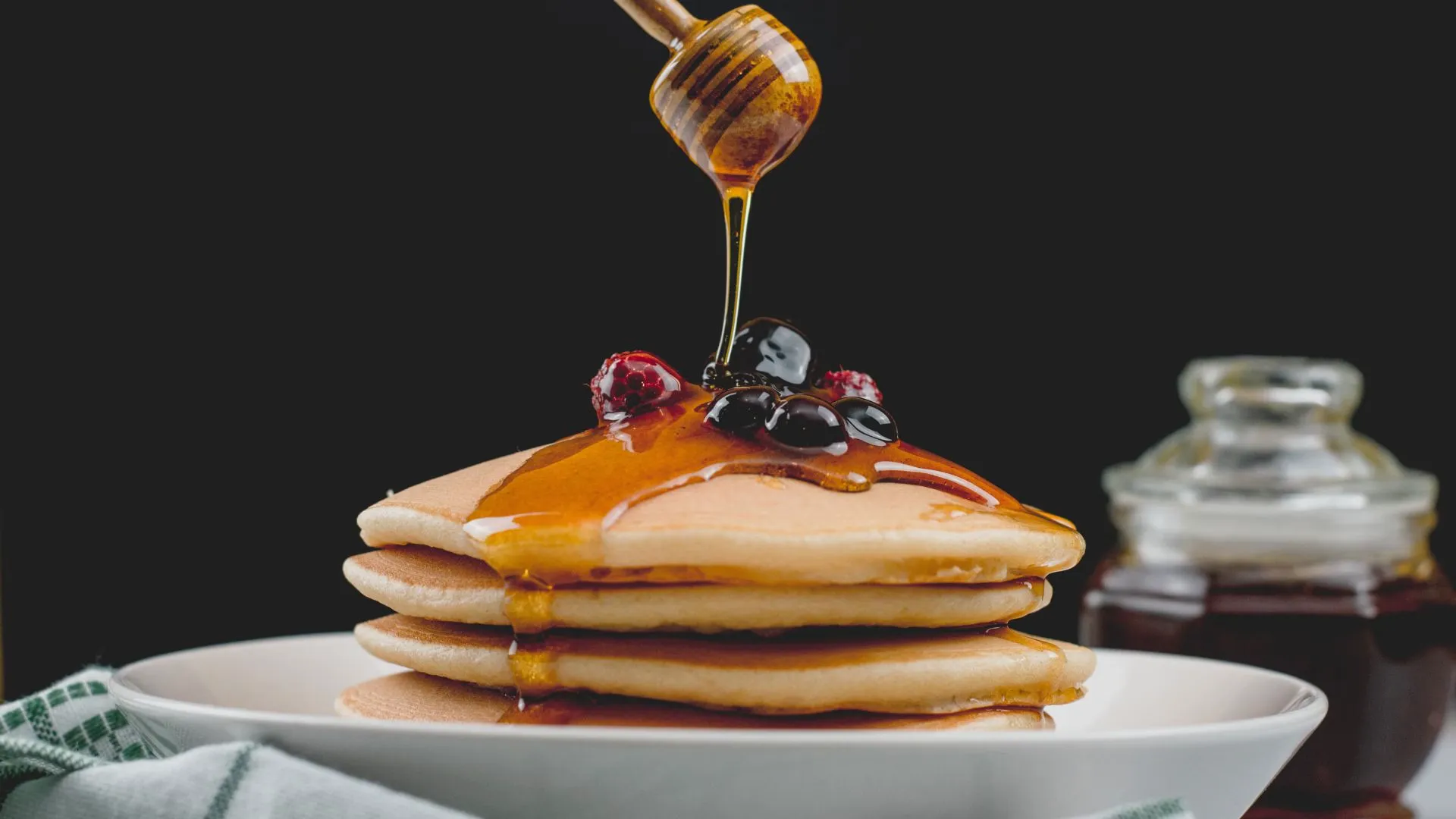 Honey dripping from a wooden dipper onto a stack of pancakes topped with berries, against a black background