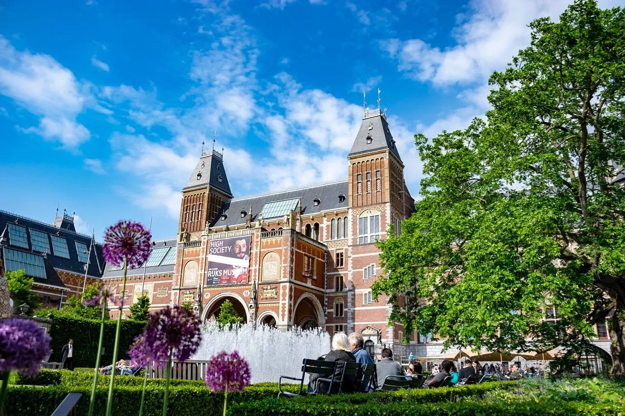 The brick towers of the Rijksmuseum in Amsterdam rise behind a fountain, green trees and purple allium flowers on a sunny day