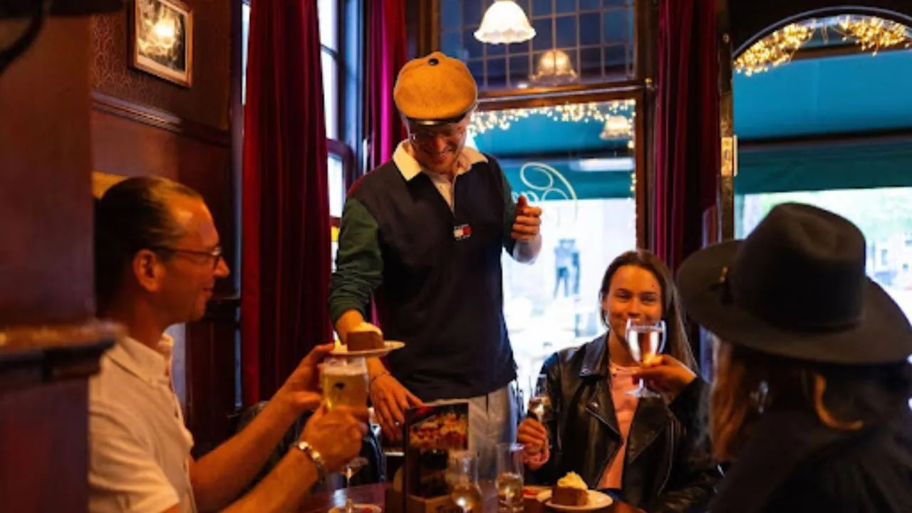 Guide in a flat cap serves a plate of cake to a smiling group seated with beers in a cozy wood-paneled Amsterdam pub