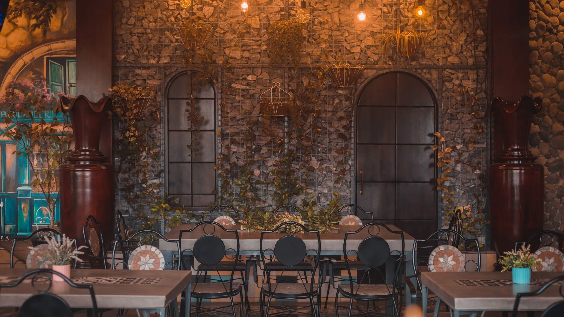 Restaurant interior with a rough stone wall, climbing plants, metal chairs and wooden tables under warm hanging lights