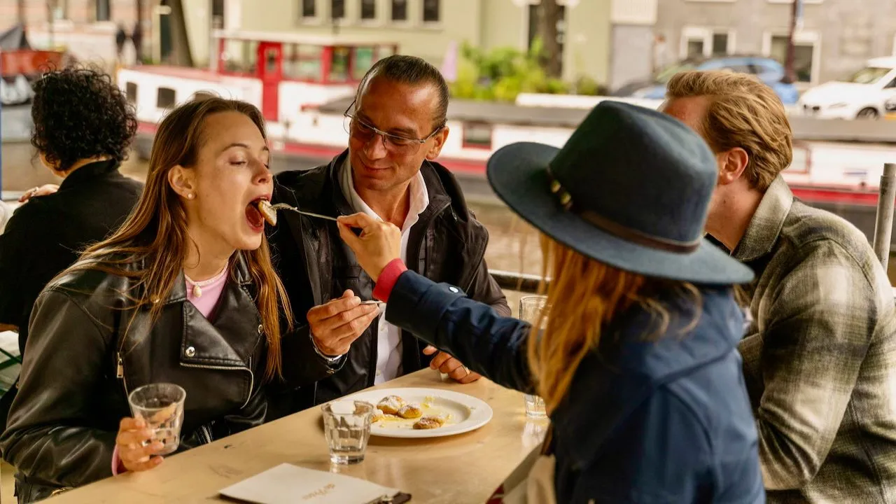 Woman in a blue hat feeds a laughing guest a bite from a fork at a canal-side table in Amsterdam while two men look on smiling