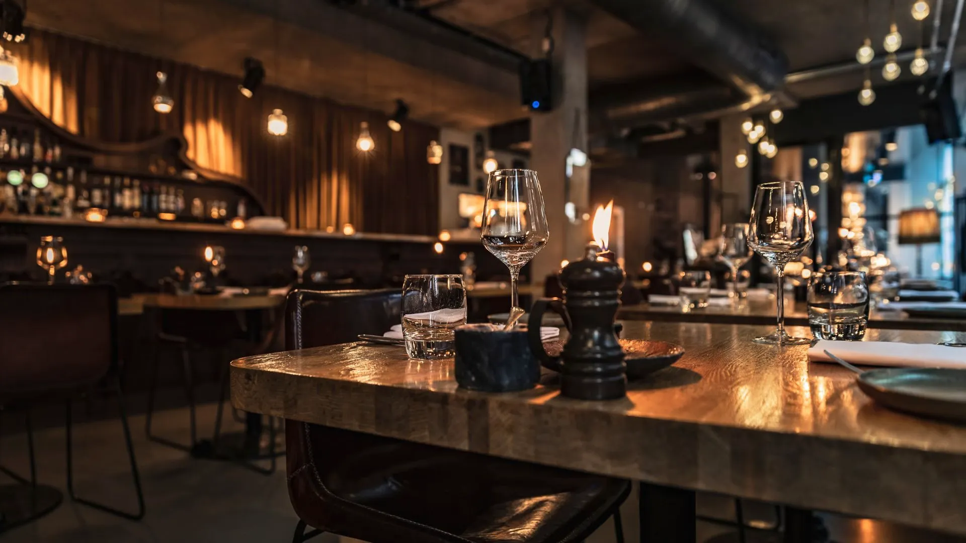 Moody restaurant interior with wine glasses, a candle and a pepper mill on a thick wooden table, warm lights glowing behind