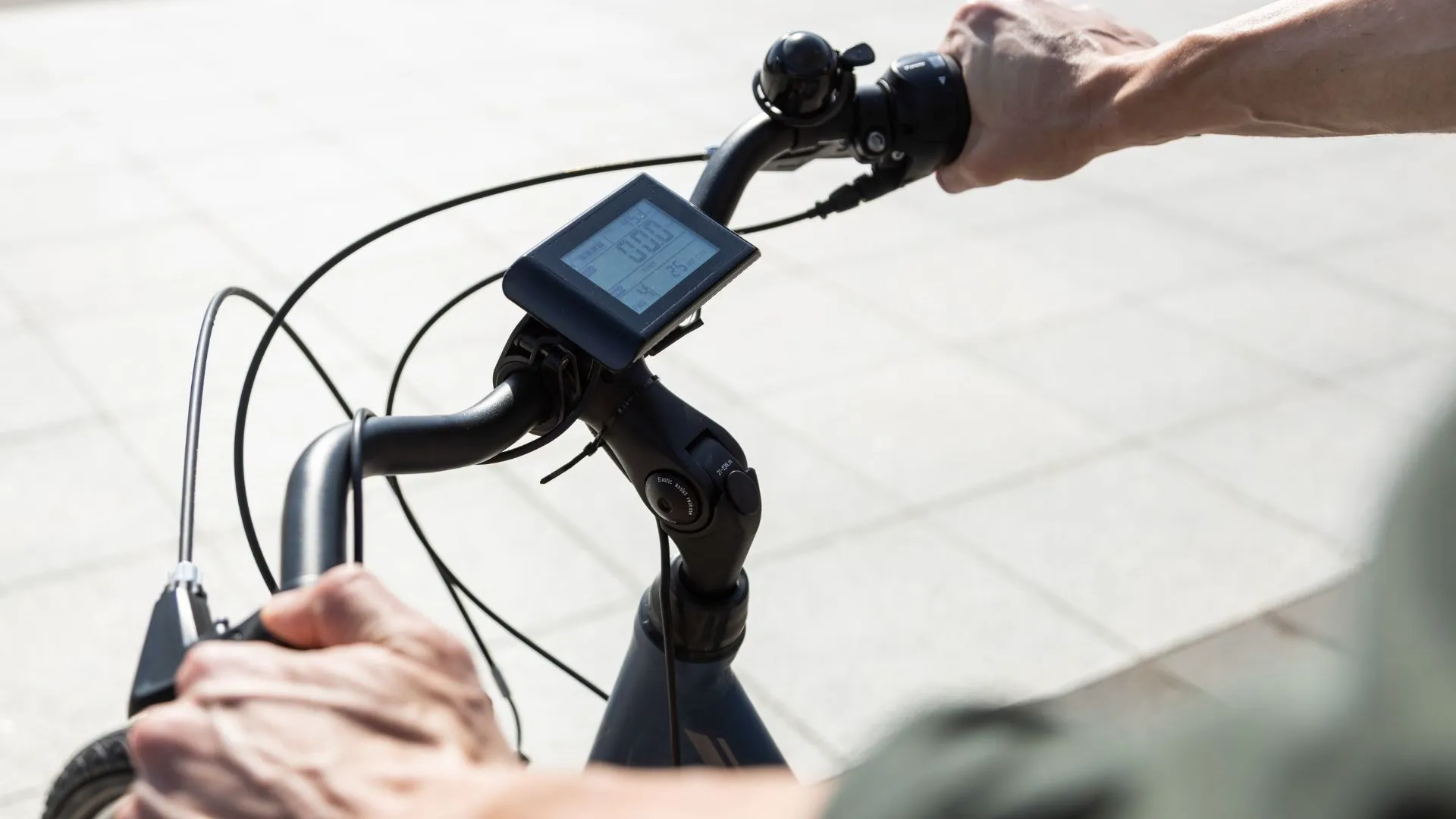 Close-up of hands on an e-bike handlebar with a small digital display mounted in the center